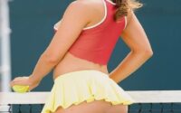 Smiling woman in a red tennis top and yellow skirt stands at the net, holding a tennis ball in front of her on a court.
